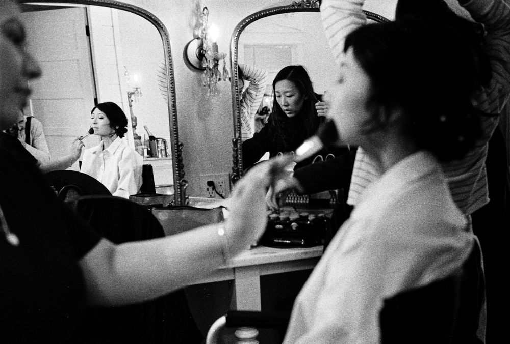 Black and white photo of a bride getting makeup applied, reflected in an ornate mirror in a vintage dressing room.