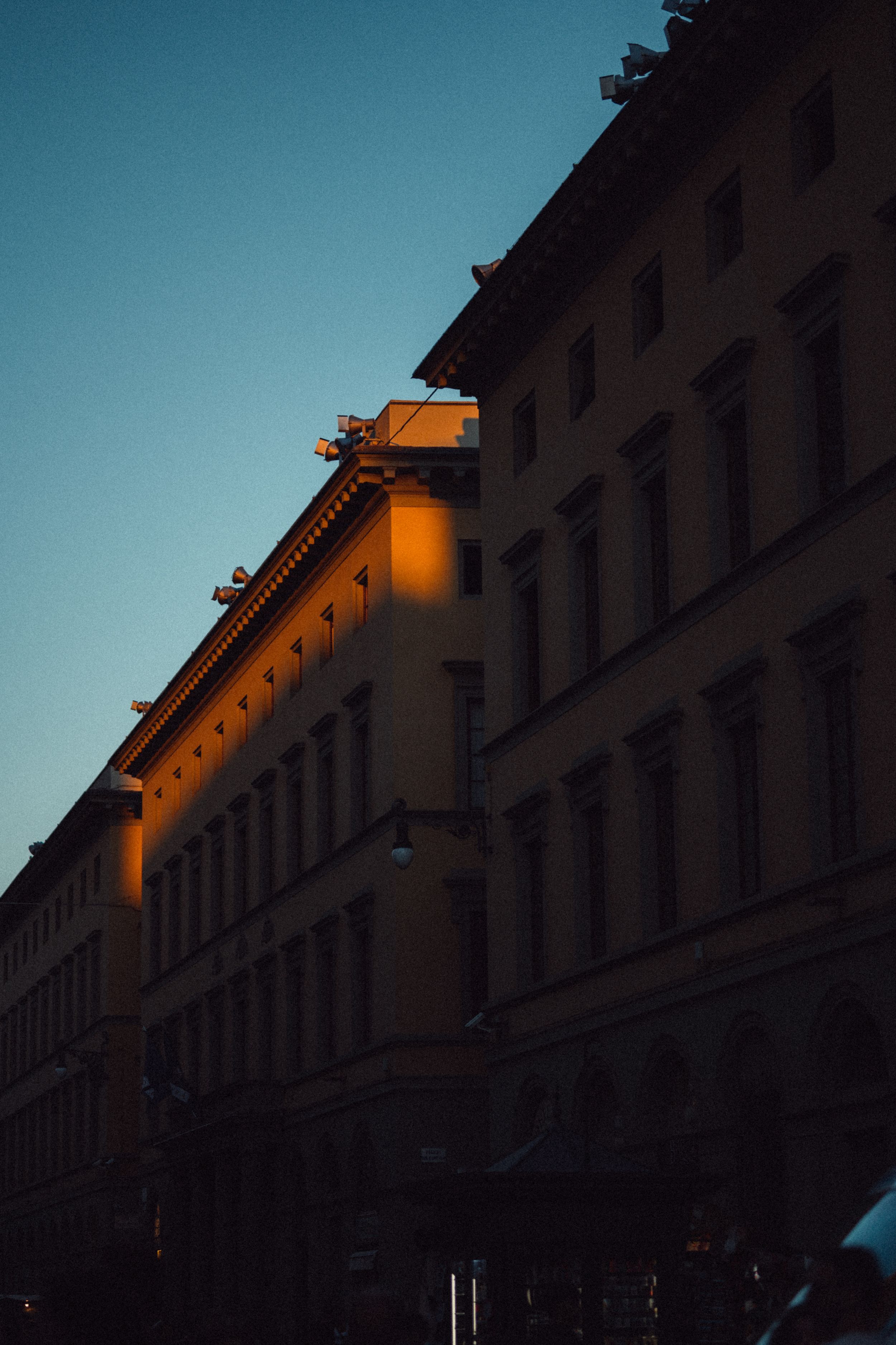 Warm evening light illuminates the top corner of a historic European apartment building against a deep blue sky.