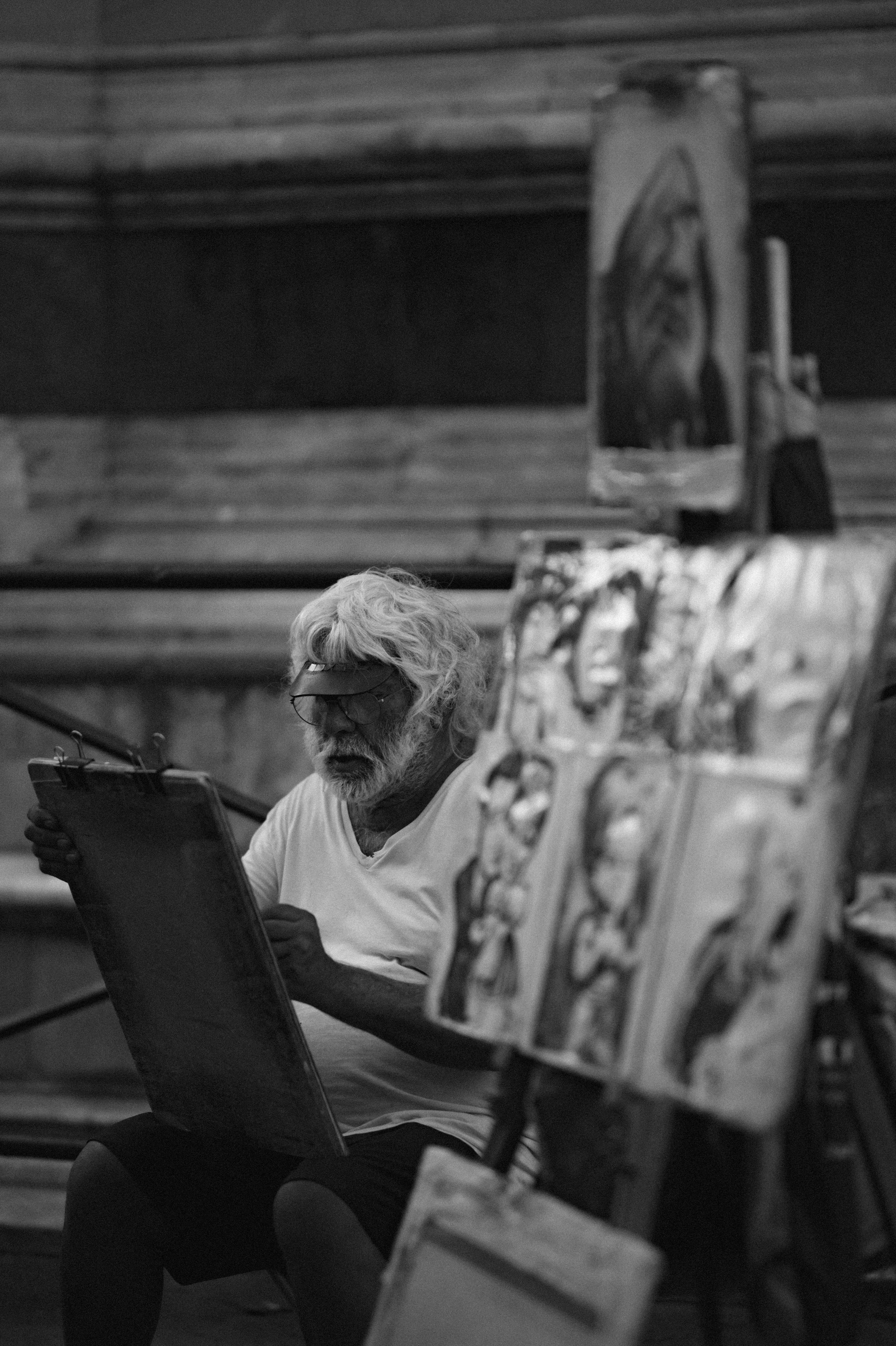 Elderly artist in white shirt sketches on clipboard while sitting next to displayed artwork in black and white scene.