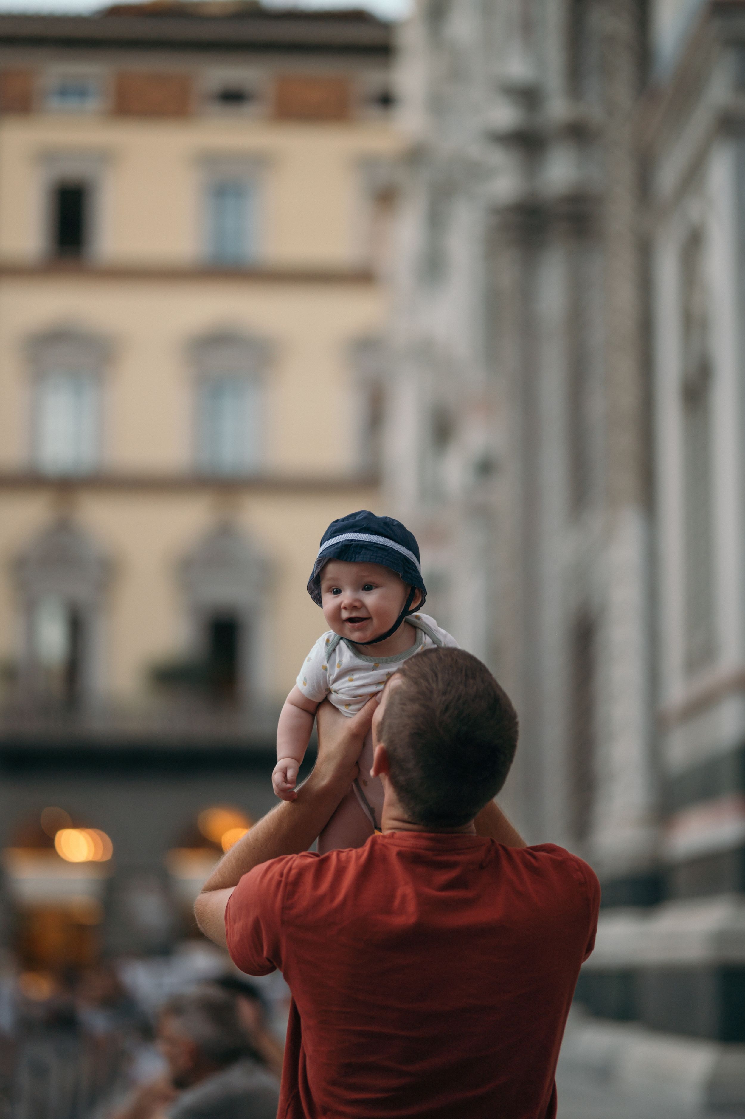 A parent in a red shirt lifts up a smiling baby wearing a striped hat and scarf in an urban setting at dusk.
