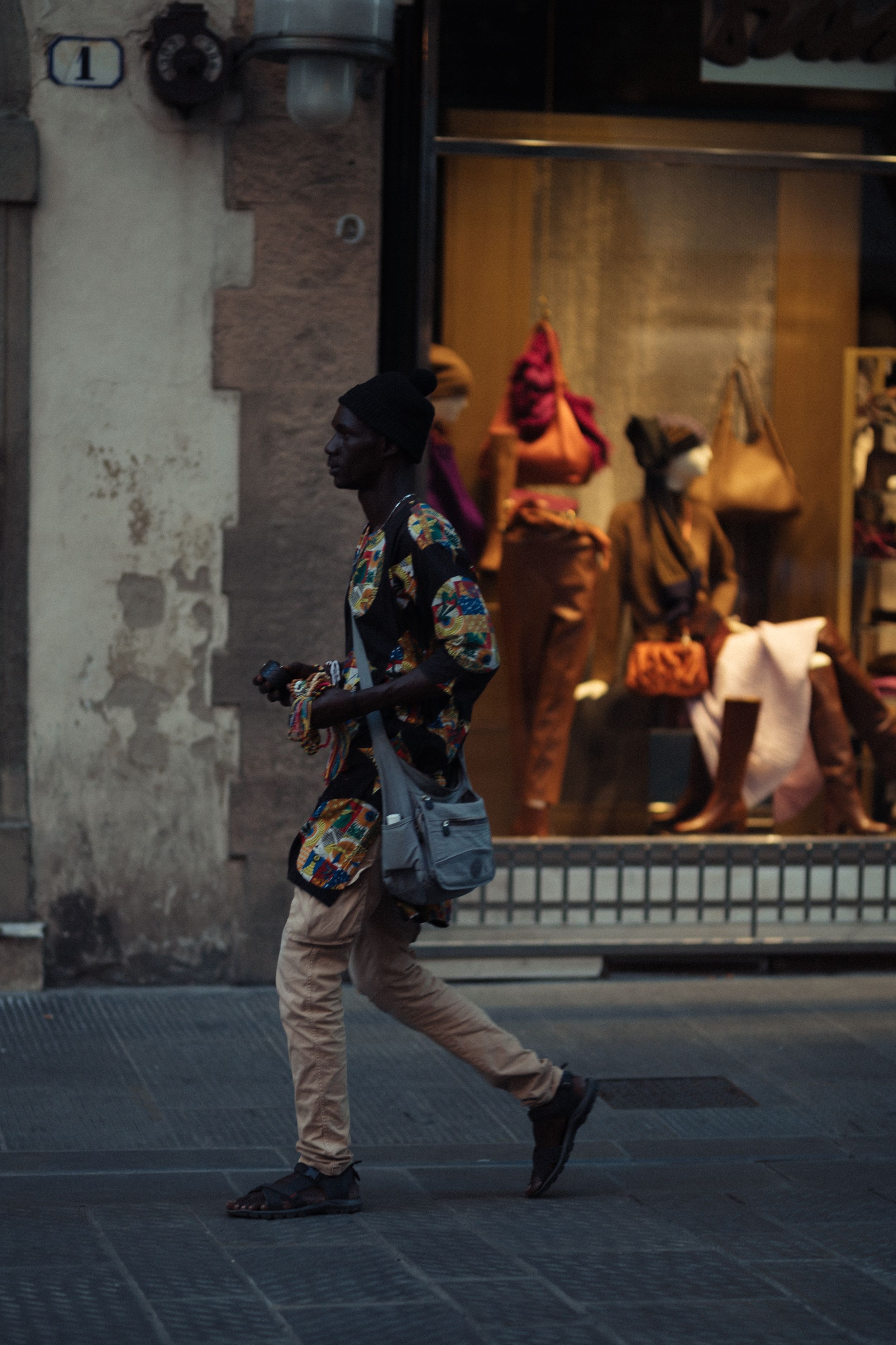 Person wearing dark hoodie and tan pants performs dynamic dance moves against urban storefront backdrop.