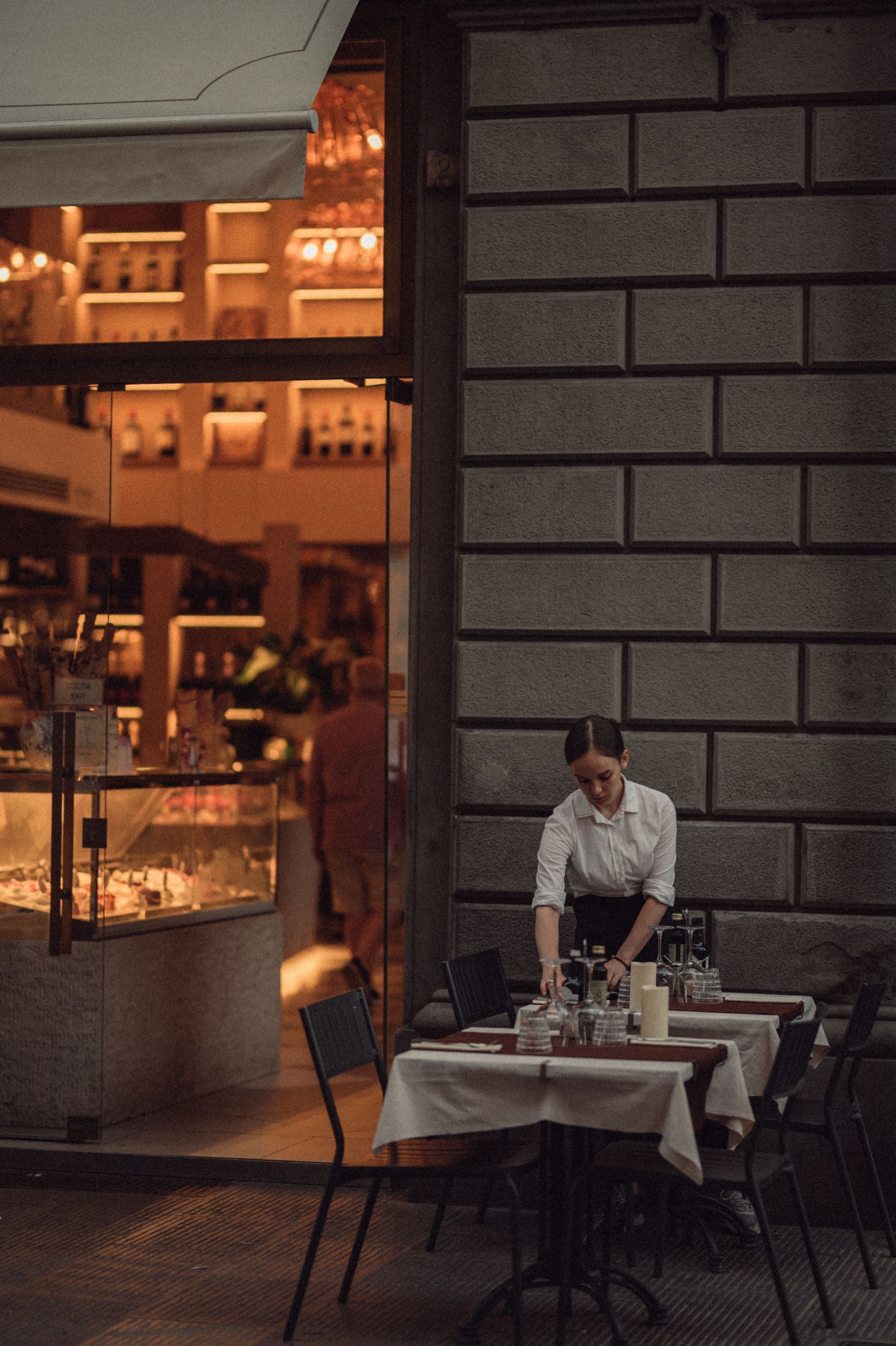Restaurant staff sets up outdoor dining tables at dusk with warm glowing lights from the bar visible inside.