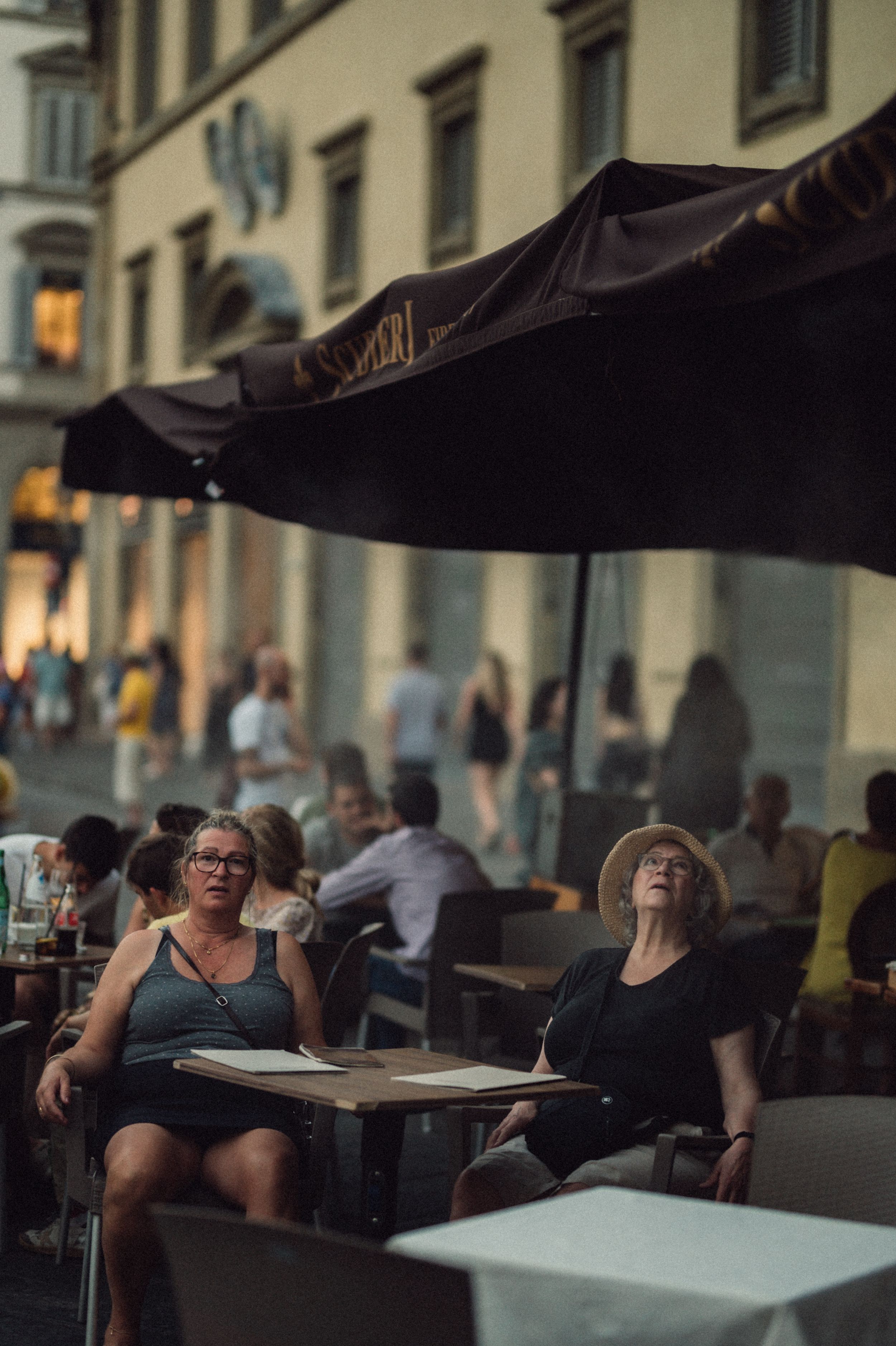 People relax at outdoor cafe tables under black umbrellas on a warm evening in a European city street.