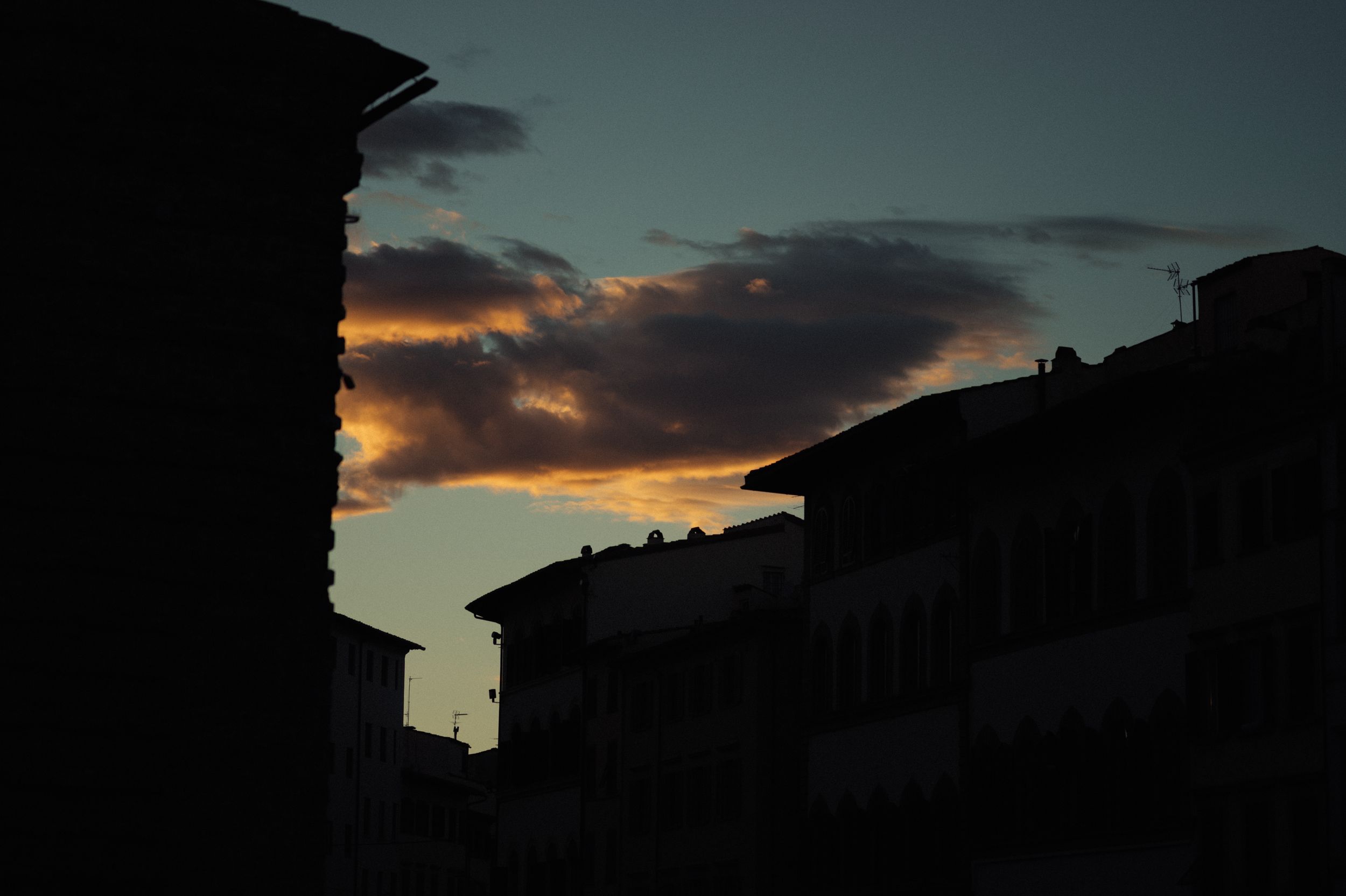Dramatic sunset sky with dark silhouettes of buildings and clouds casting an orange glow across the horizon.