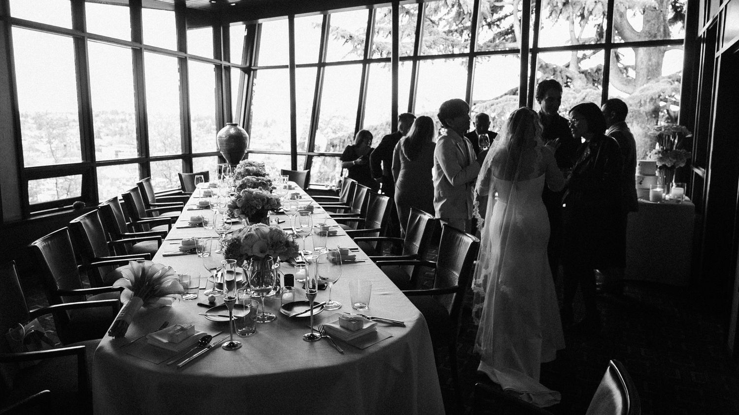 Black and white photograph of a formal dining table setting with guests gathering near windows at an elegant event.