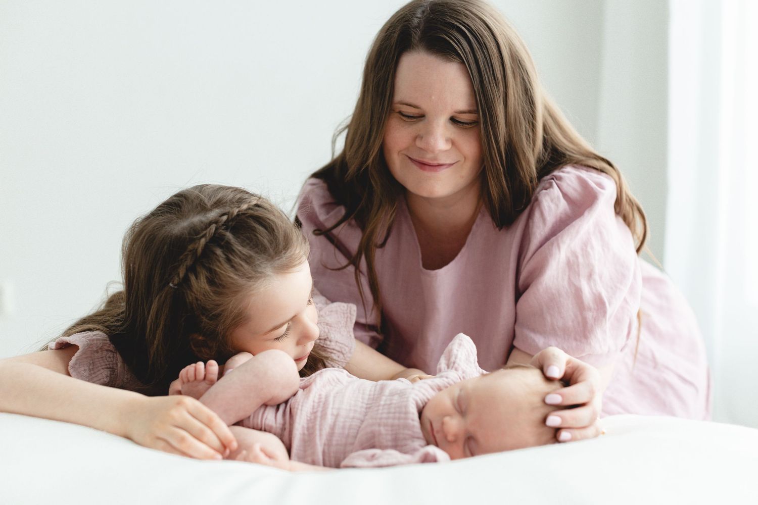 Tyttövauva uinuu beanbagin päällä valoisassa studiossa, tuore isosisko silittää vauvaa. Ylpeä äiti taustalla. Koko perheen newborn-kuvaus.