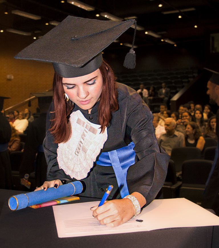 Joyce graduating in cap and gown signs their diploma at graduation ceremony.