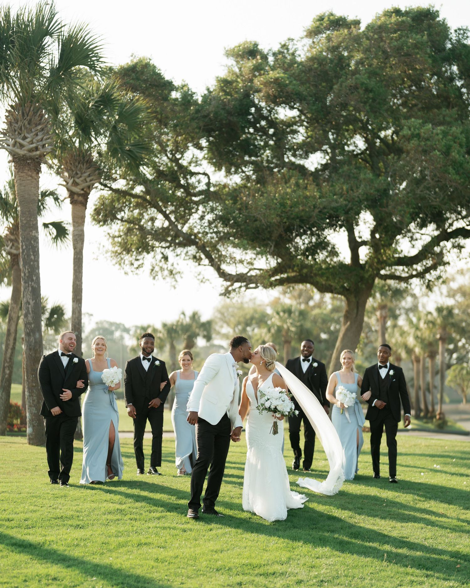 Wedding party walking on lush green lawn with palm trees and oak trees in background.