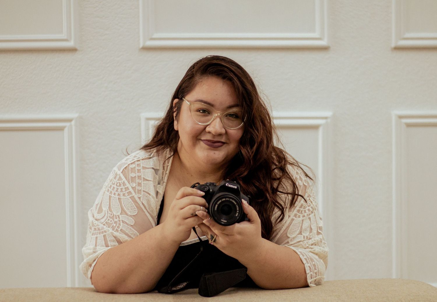 Photographer in white lace top leans on table while holding professional camera and smiling warmly.