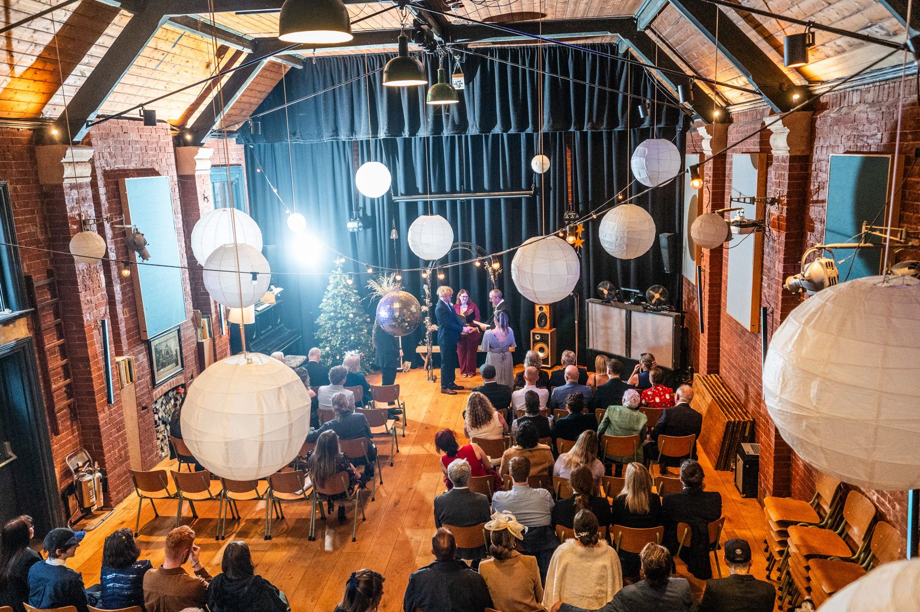 A wedding ceremony venue with exposed wooden beams and hanging white paper lanterns illuminating guests seated in rows.
