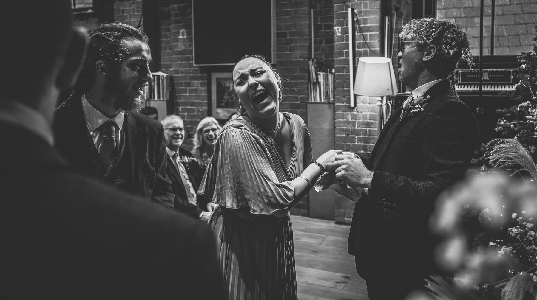 Wedding guests mingle in a rustic brick-walled space during a lively evening reception celebration.