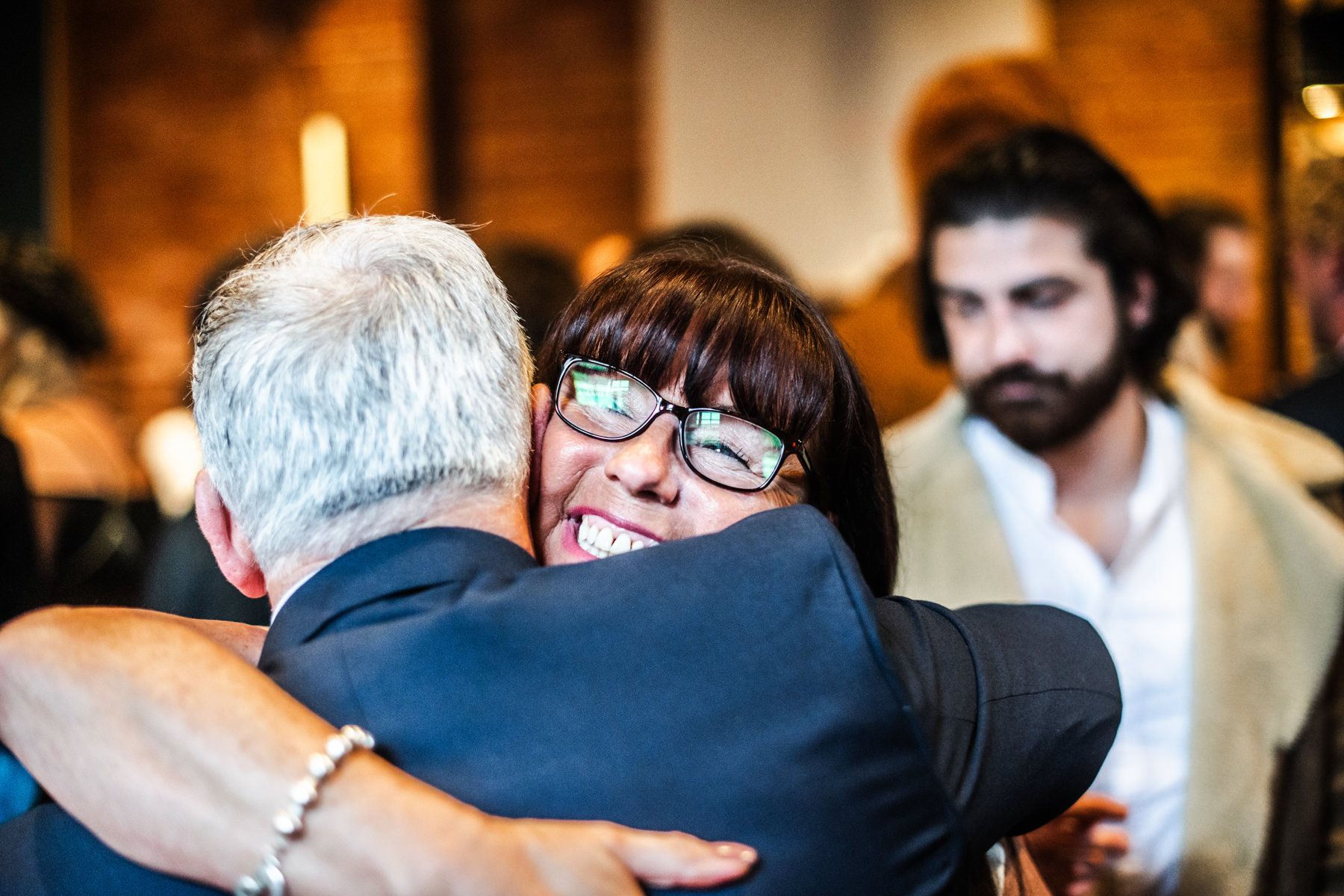 Two people embrace in a warm hug at an indoor event with warm lighting in the background.