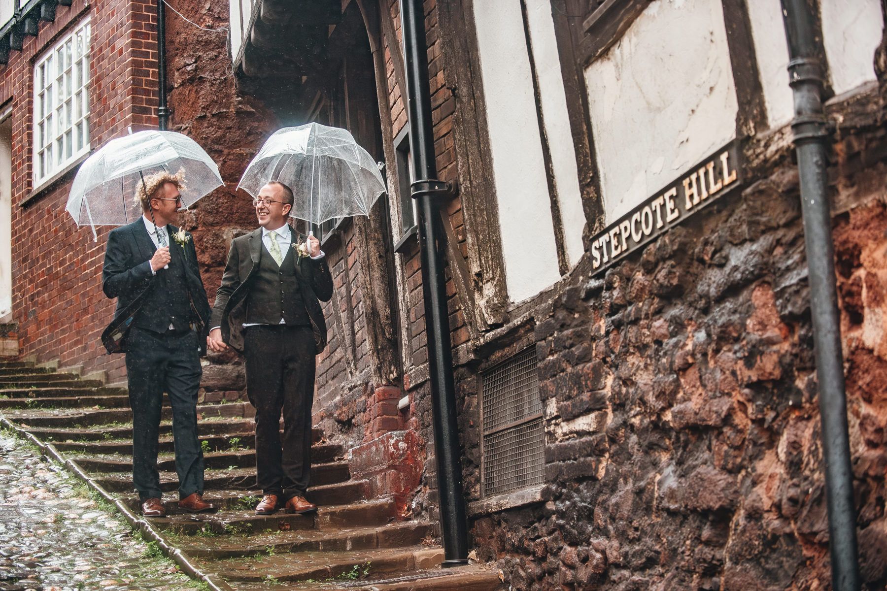 Two figures in dark suits hold white umbrellas while descending stone steps by a red brick building in the rain.
