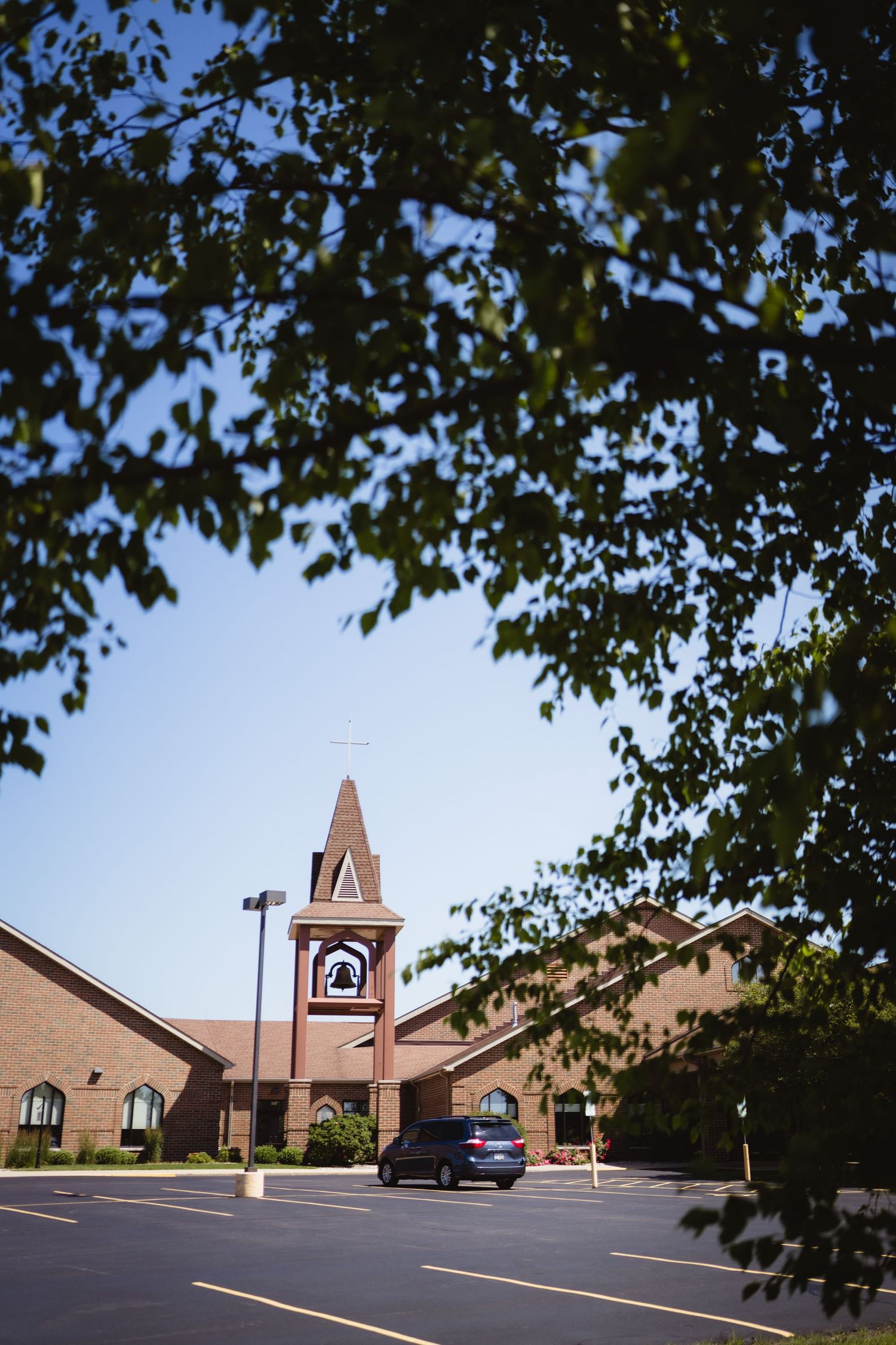 A brick church building with a bell tower and steeple is framed by green tree branches in a parking lot setting.