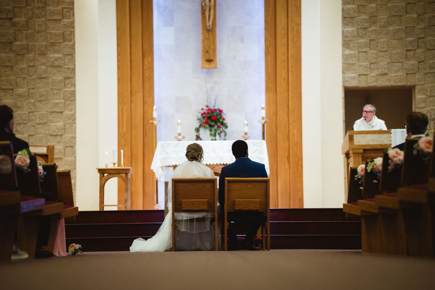 A couple sits at the altar during a traditional church wedding ceremony with wooden pews and a crucifix visible.