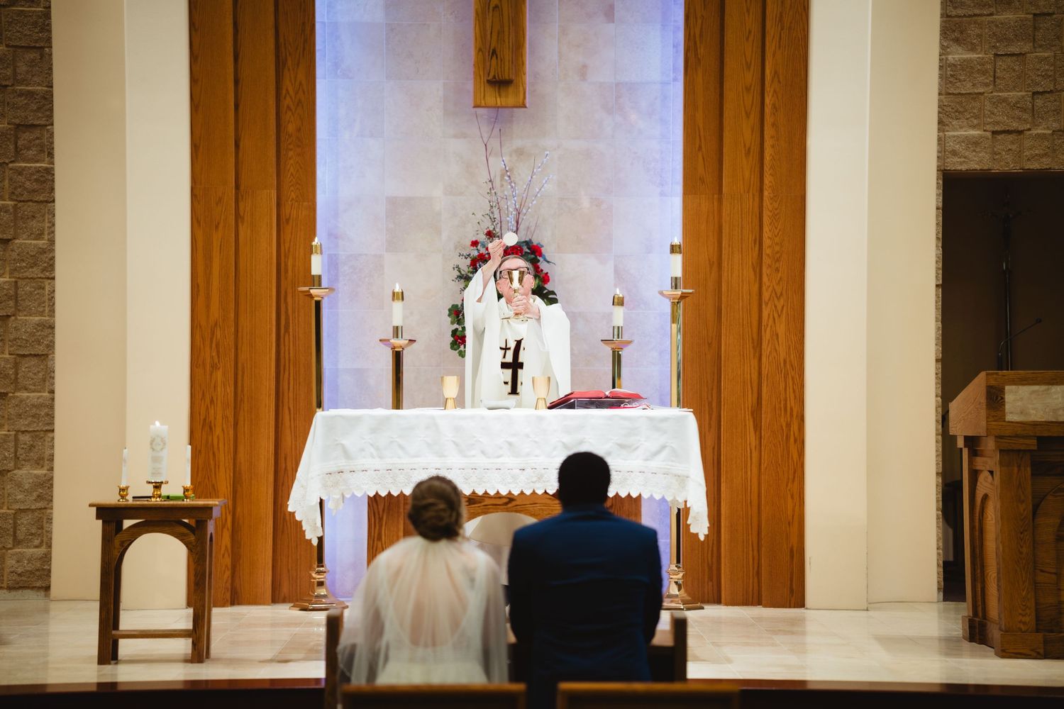 A couple sits before an altar during their wedding ceremony in a church with white walls and stone accents.