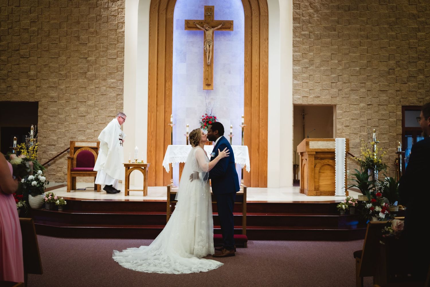 A couple shares their first kiss as newlyweds at the altar in a church sanctuary with a large wooden crucifix on the wall.