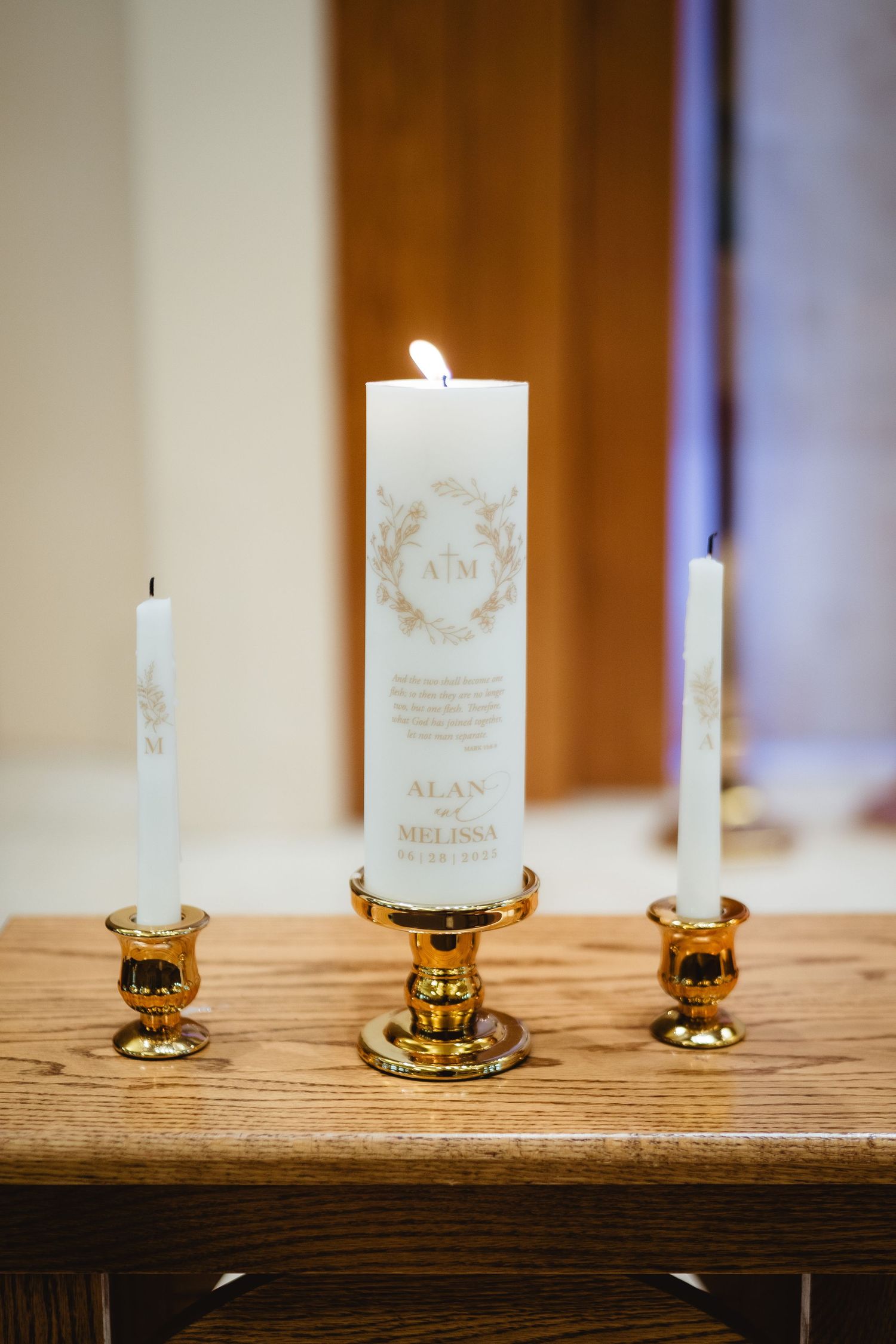 Three white pillar candles in brass holders sit atop a rustic wooden table at a wedding reception.