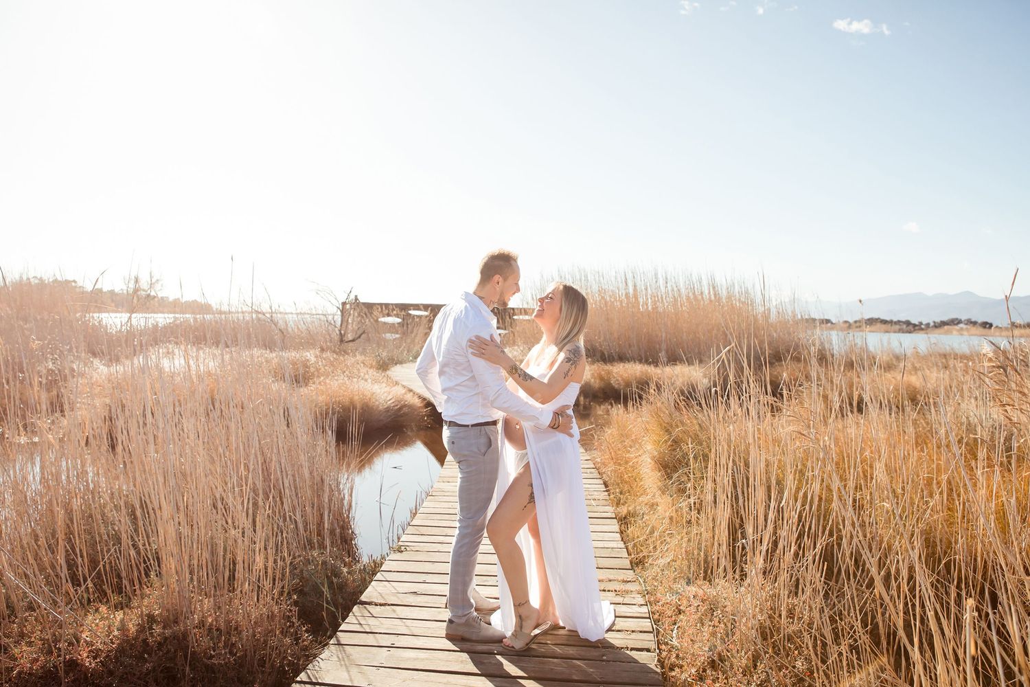 séance photo extérieur grossesse couple photographe Couple embracing on wooden boardwalk surrounded by tall coastal grasses at sunset.