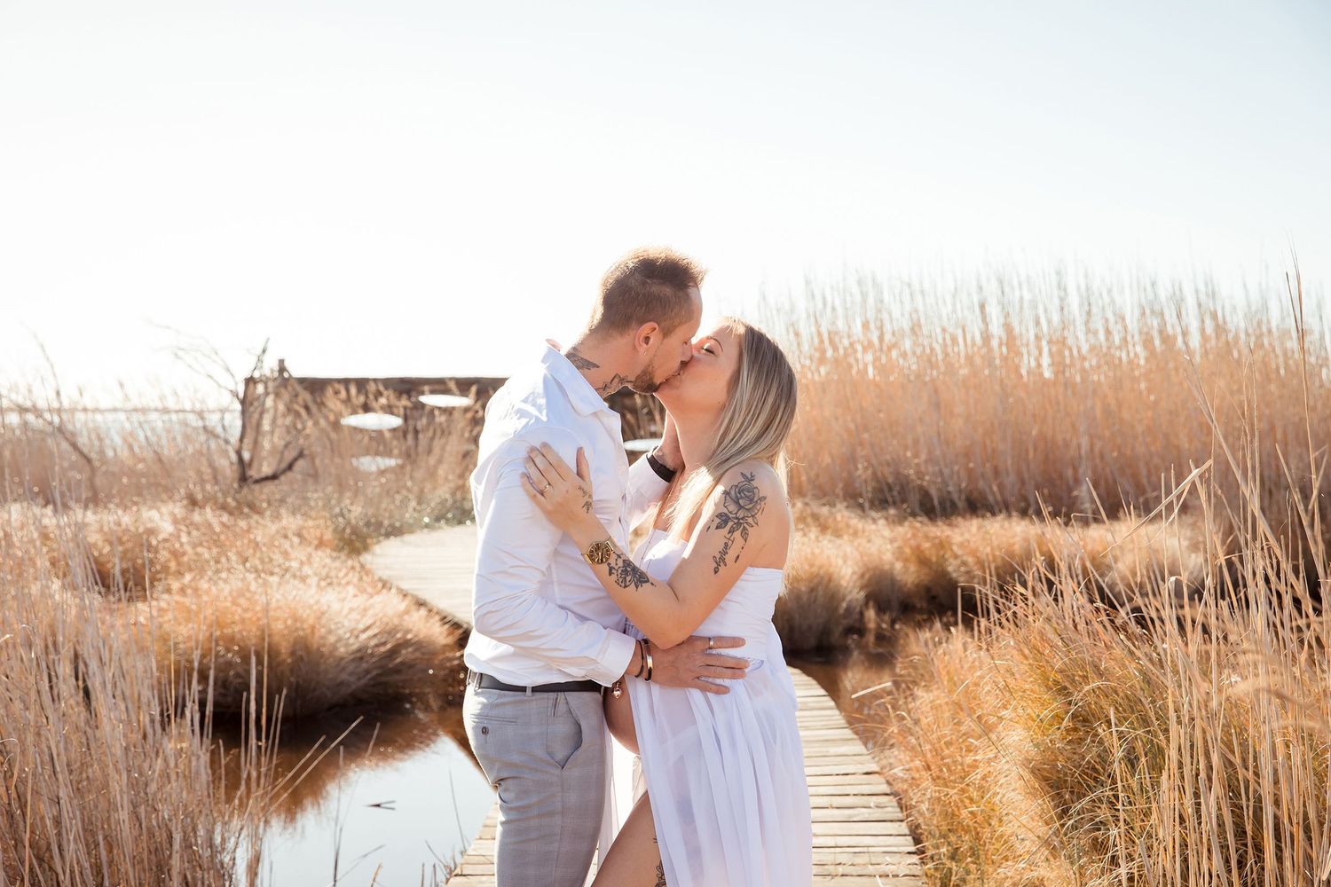 séance photo extérieur grossesse couple photographe Couple embracing on wooden boardwalk surrounded by tall coastal grasses at sunset.