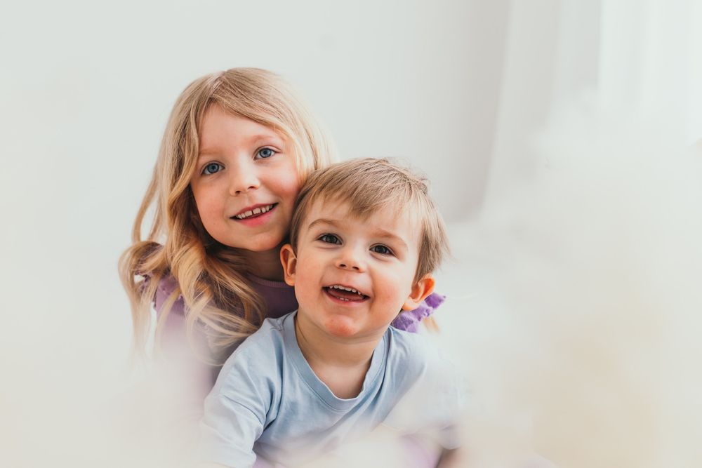 Two happy young children, a blonde girl and a toddler boy, smiling together in a bright white room.