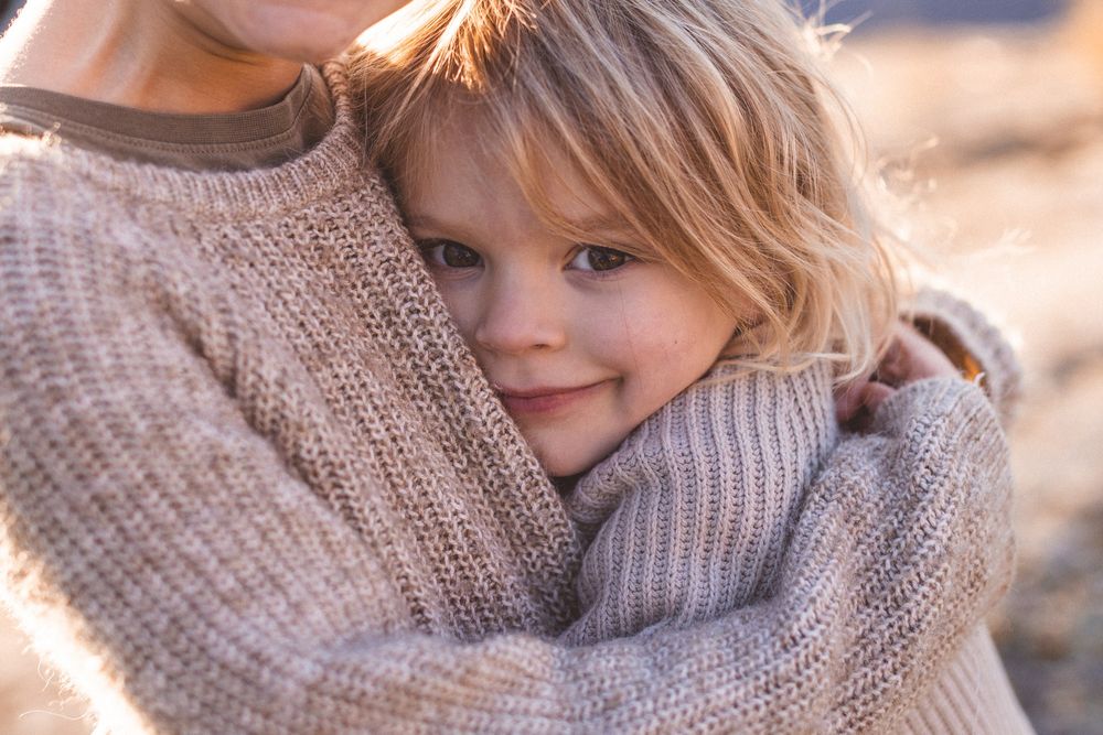 A young smiling child hugs an adult warmly, both wearing cozy knit sweaters in soft neutral tones outdoors.
