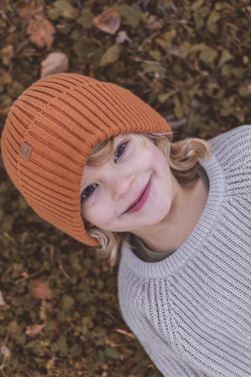 Smiling child wearing an orange knit beanie and gray sweater, sitting among autumn leaves outdoors.