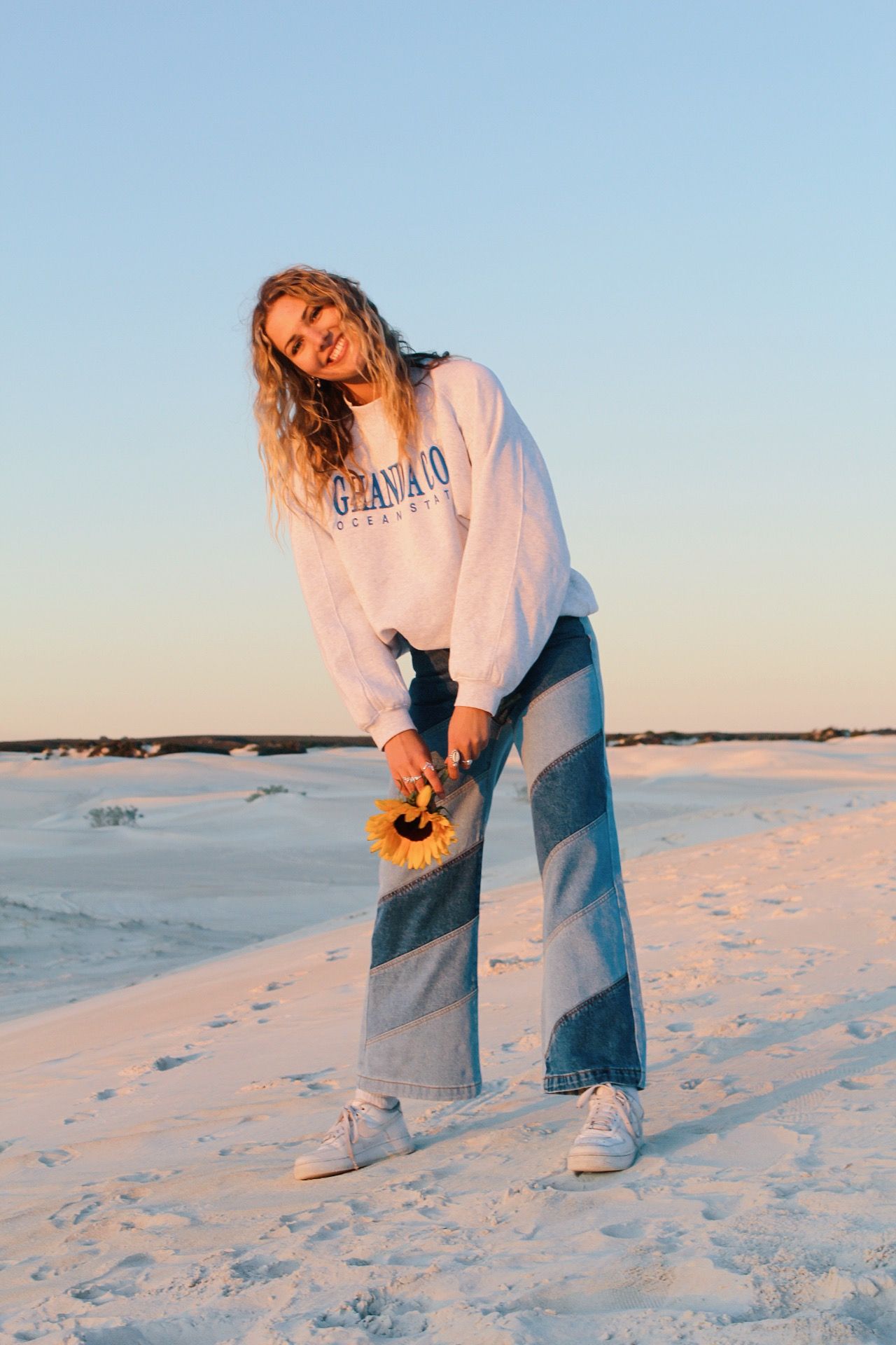 Person in casual wear poses playfully on white sandy beach at sunset.