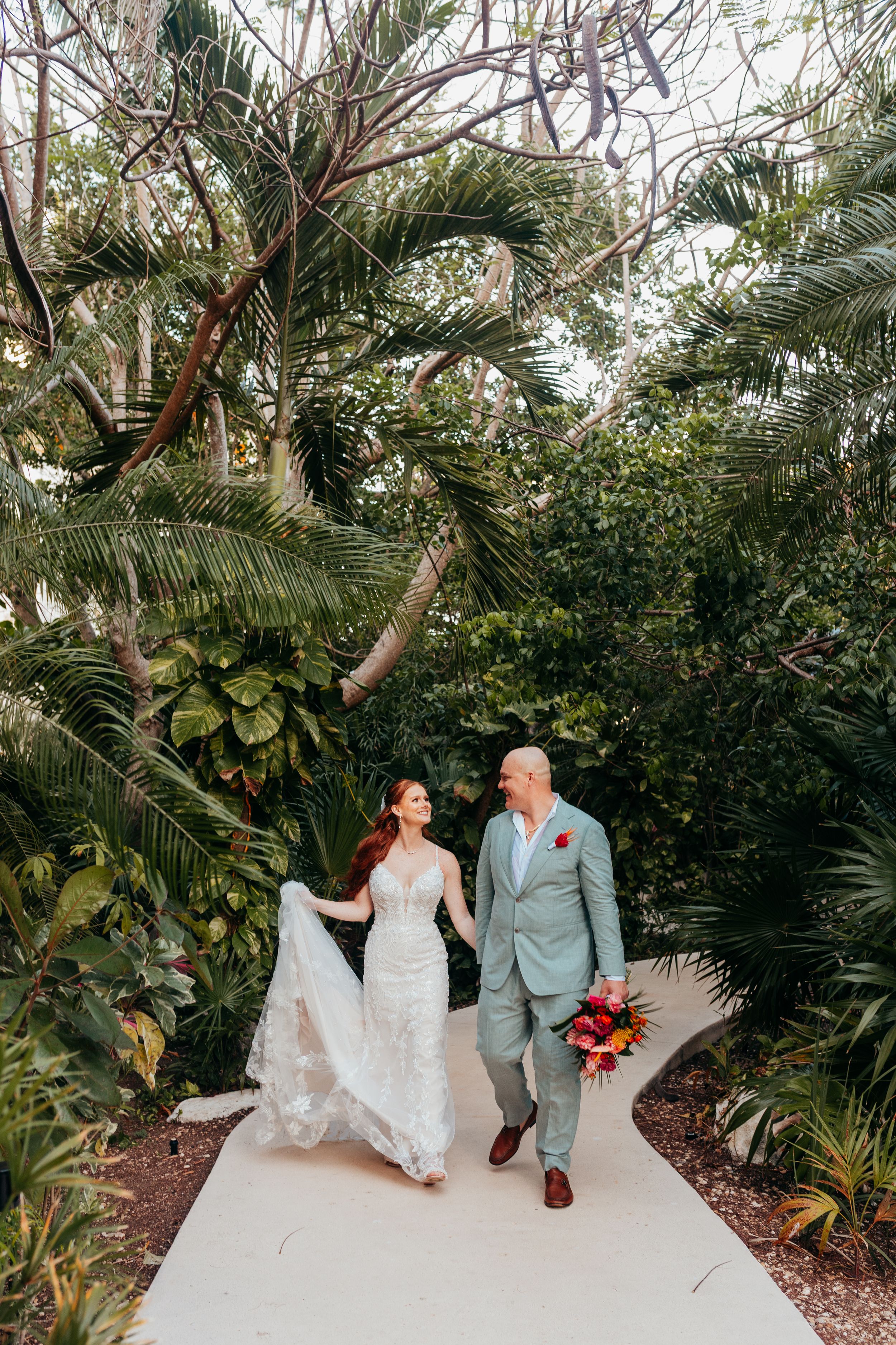 Wedding couple walks hand in hand along tropical garden path surrounded by lush palm trees and greenery.