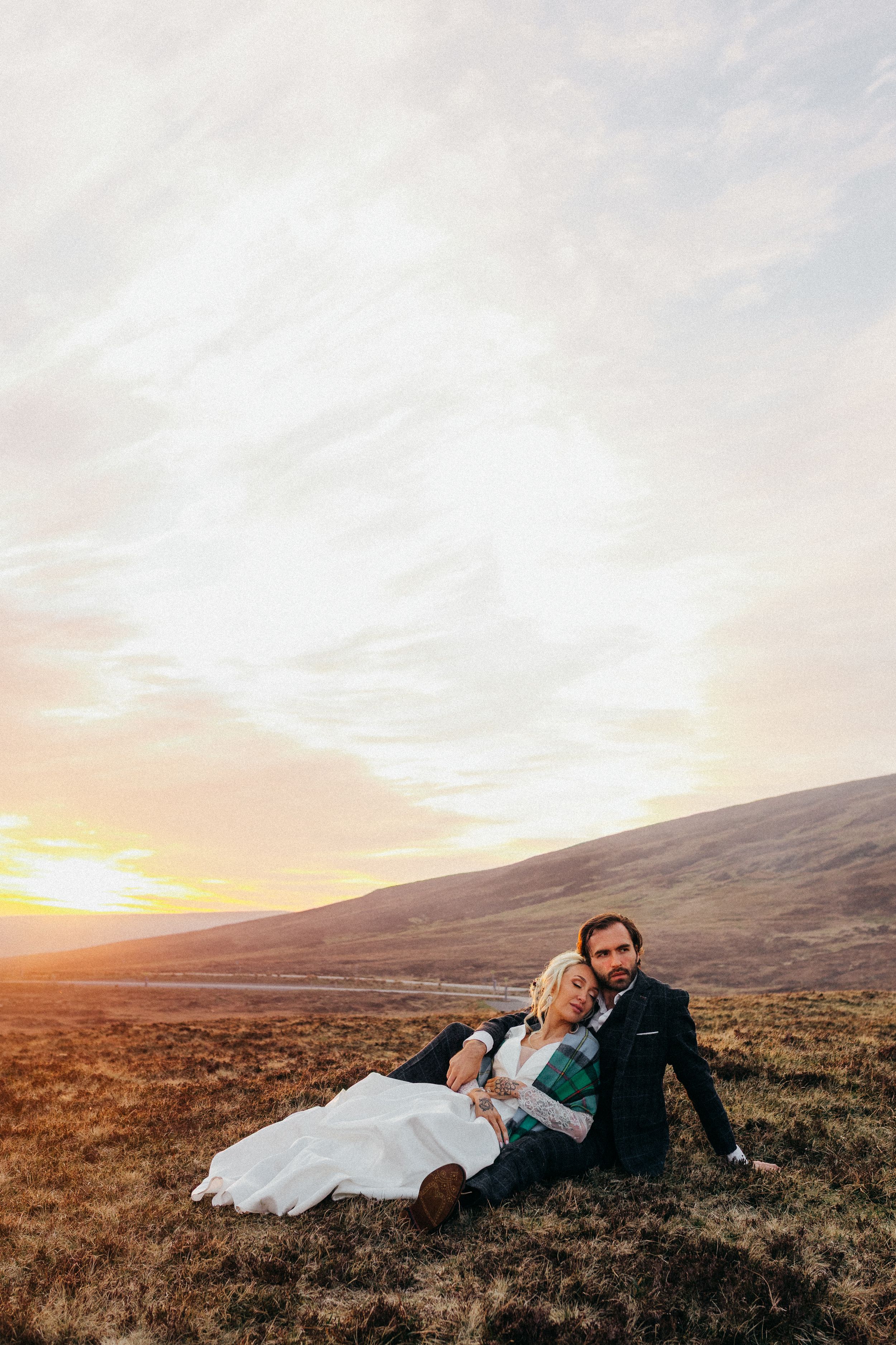 Two people sit together on a hillside at sunset with mountains in the background and golden light illuminating the landscape.