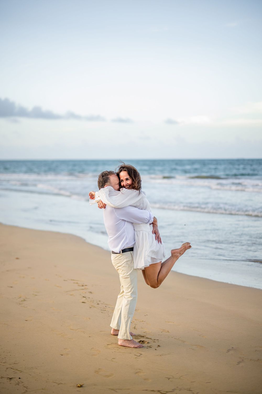 A couple enjoys playful lifts and embraces on a serene beach at sunset with ocean waves in the background.