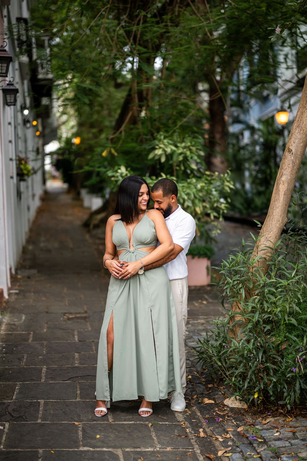 A couple walks intimately along a stone pathway surrounded by lush green foliage with dappled sunlight overhead.