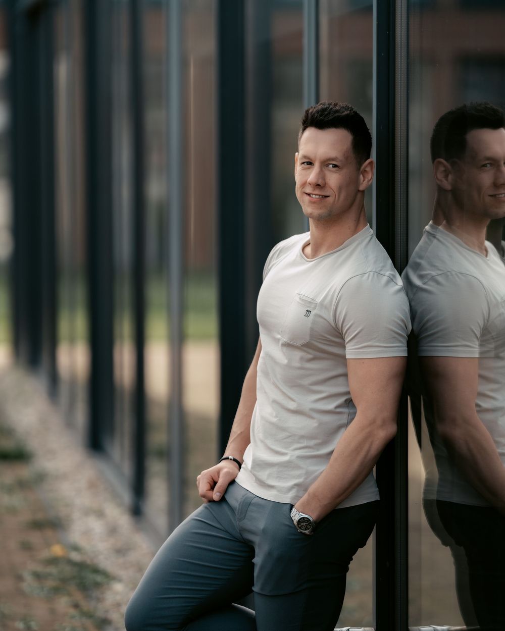 Side view of person in white t-shirt and dark pants leaning casually against reflective glass building exterior.