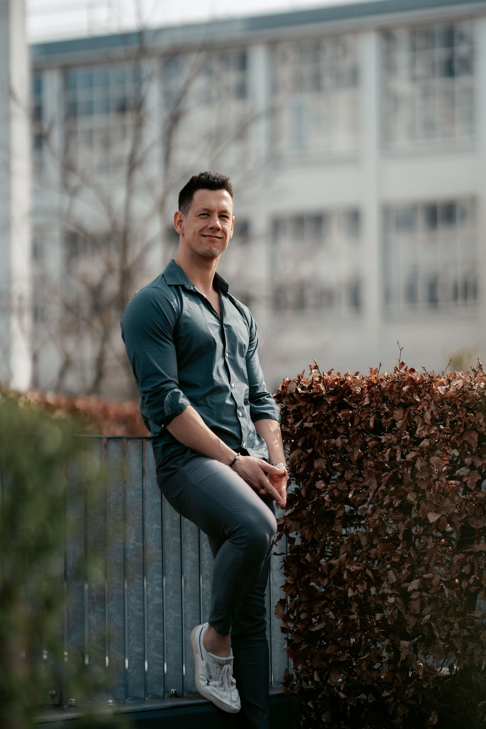Person sitting on railing wearing dark shirt and jeans against city building with hedge in foreground.