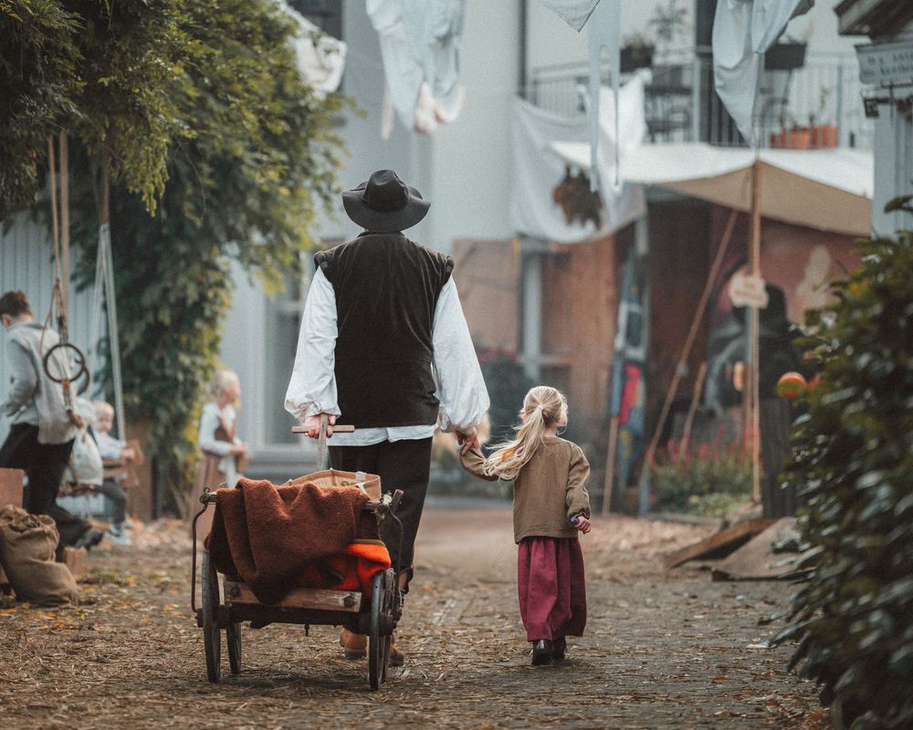 People in period costumes walking along a tree-lined path during a historical reenactment.