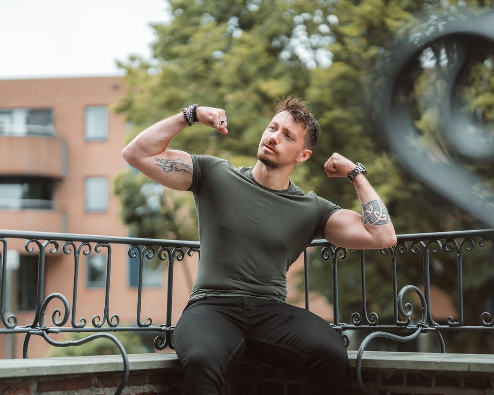 Athletic person in olive green t-shirt flexing muscles while posing against wrought iron railing outdoors.