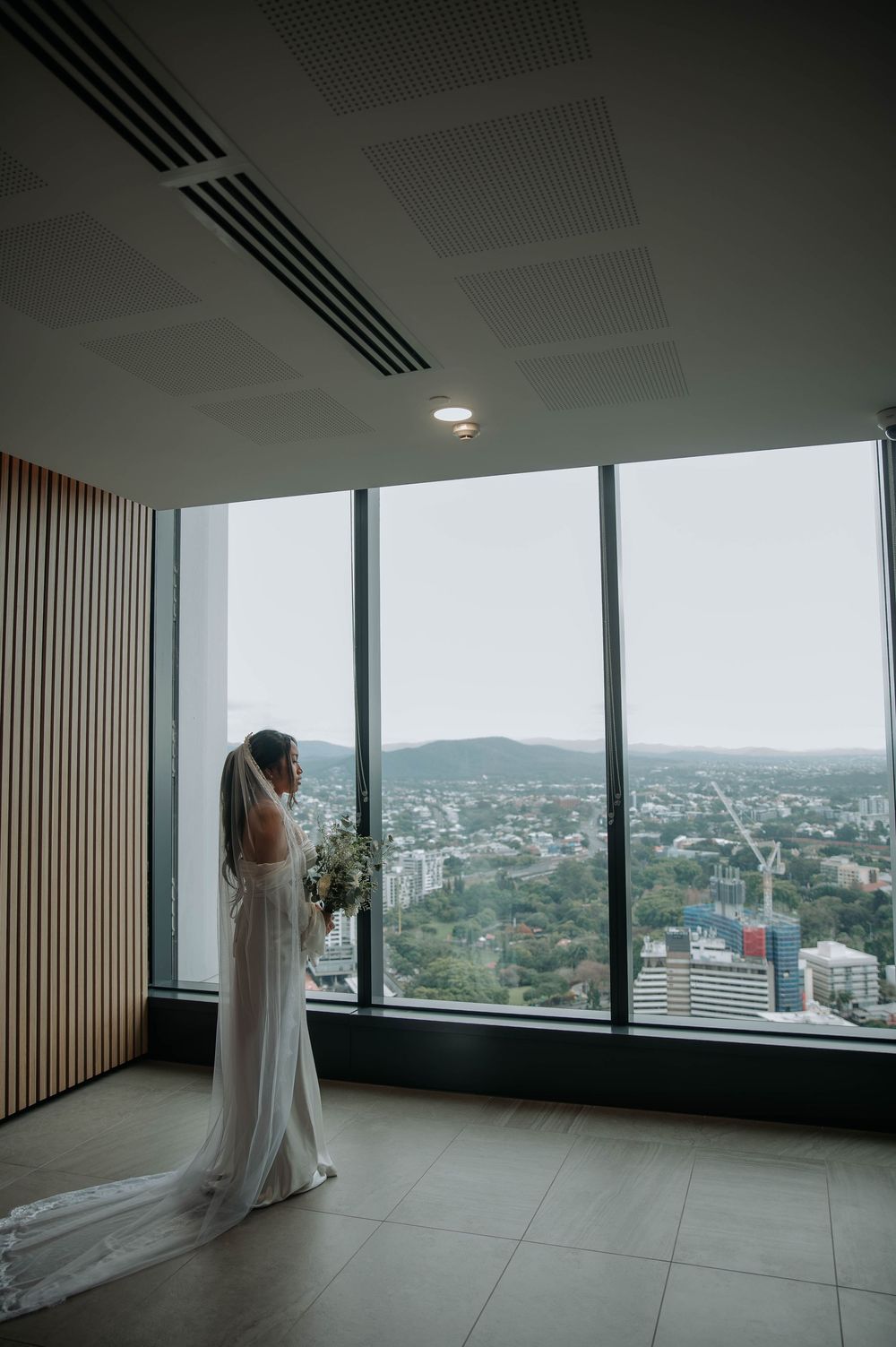 Person in flowing white dress stands by floor-to-ceiling windows overlooking a cityscape from a modern high-rise building.