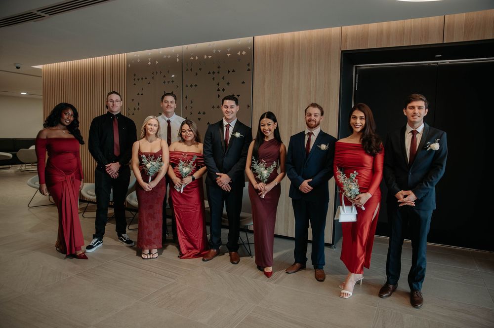 Wedding party lined up in formal attire with bridesmaids in burgundy dresses and groomsmen in dark suits against modern wall.