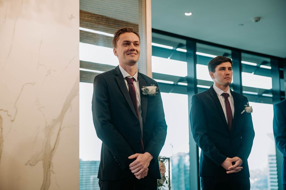 Two groomsmen in dark suits with boutonnieres stand waiting in a bright modern space with city views.