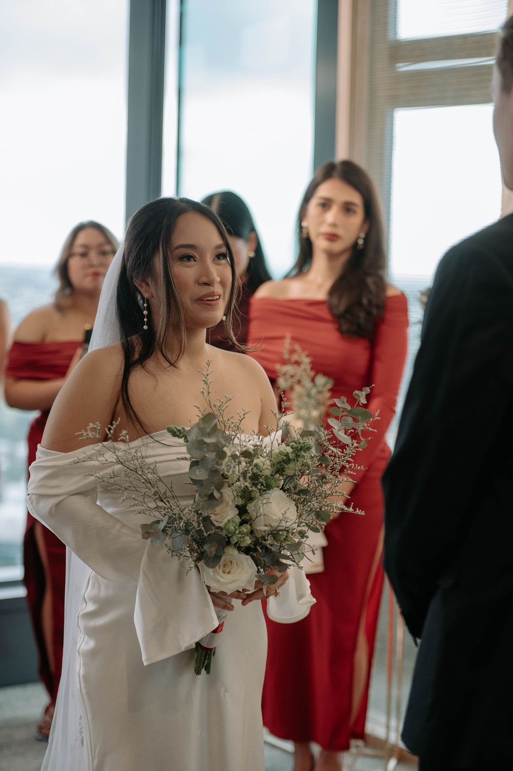 A beautiful bride in a white wedding dress holds a delicate bouquet of eucalyptus and white flowers with bridesmaids in red dresses.