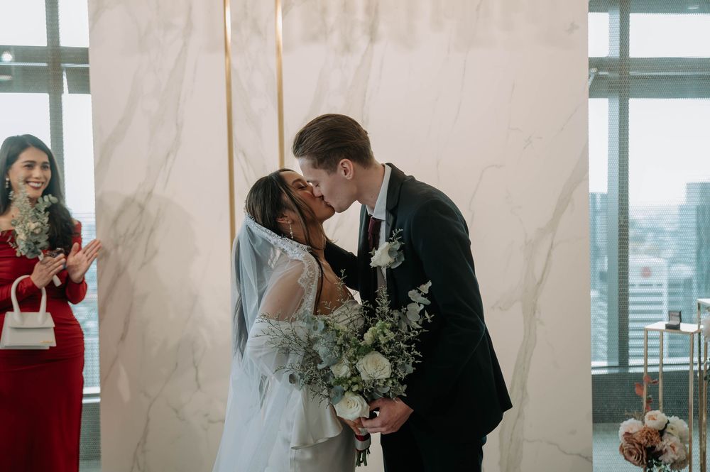 A couple shares their first kiss during an indoor wedding ceremony with a modern city backdrop through large windows.