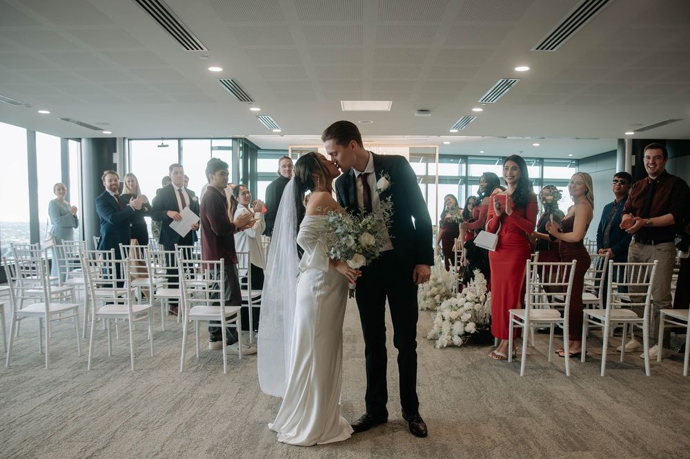 A wedding procession walks through a modern venue with white chiavari chairs lined up for the ceremony.