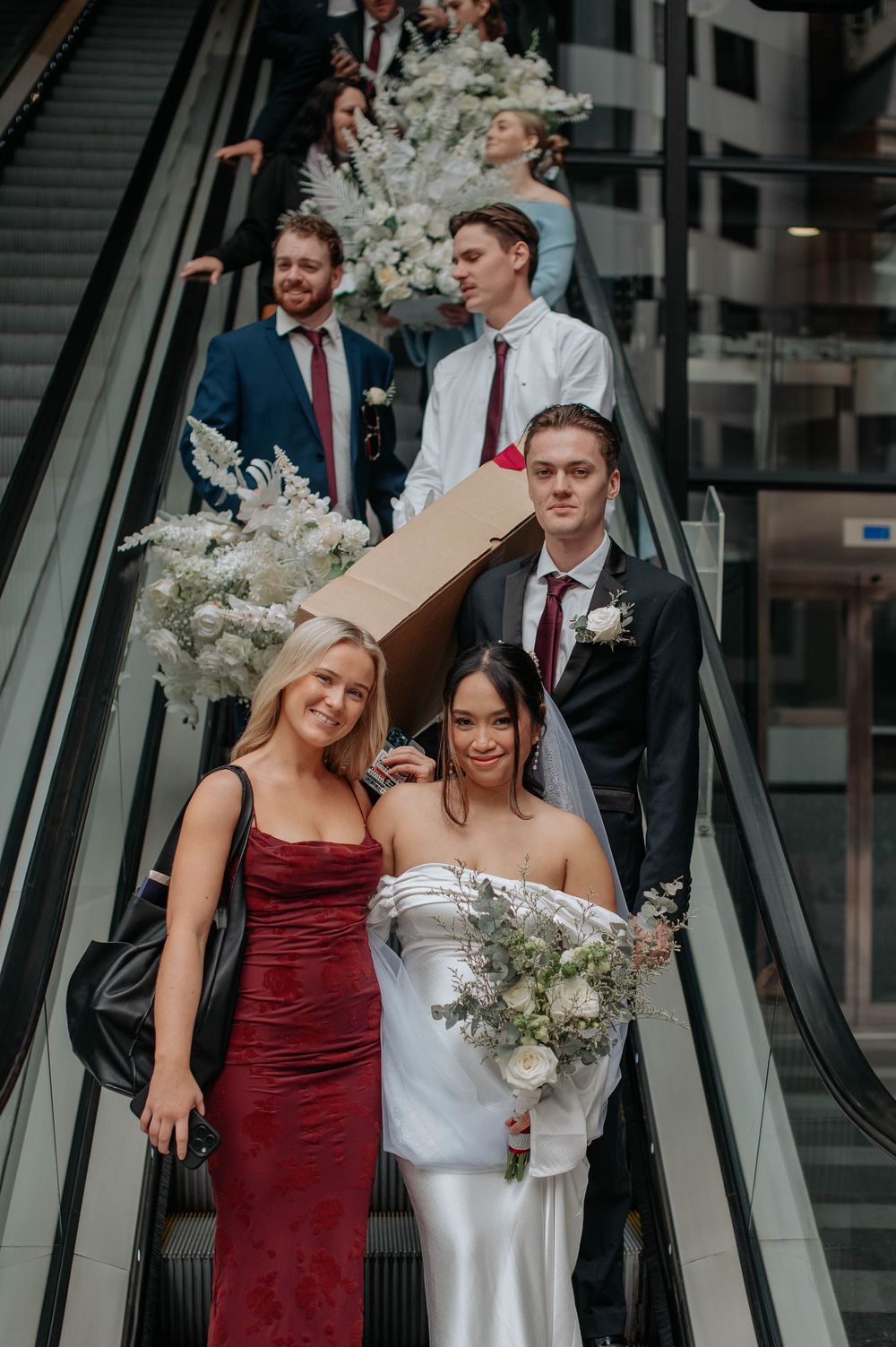 Wedding party poses on escalator decorated with white floral arrangements in modern building.