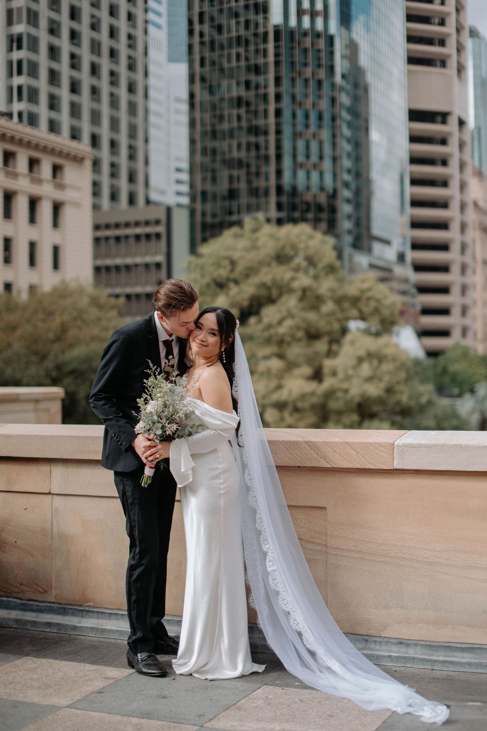 Elegant wedding couple embraces on city rooftop balcony with modern skyscrapers in background during romantic moment.