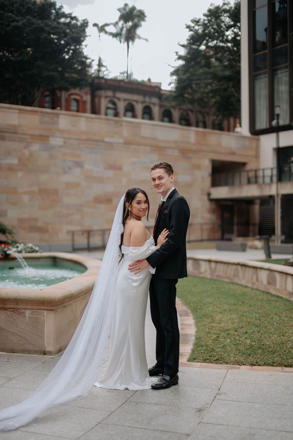 A joyful couple shares a tender moment on their wedding day against classic stone architecture with palm trees.