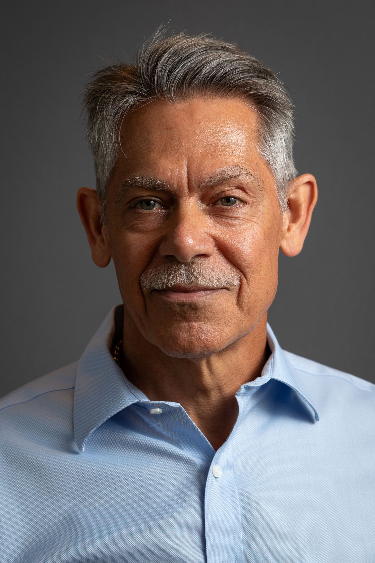 Professional studio portrait of a mature man with gray hair wearing a light blue button-down shirt against dark background.