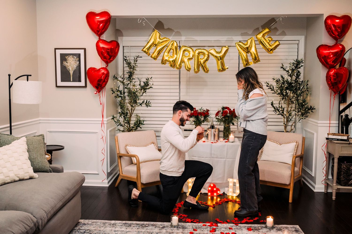 A romantic marriage proposal scene with gold 'MARRY ME' balloon letters and red heart balloons decorating a cozy living room.
