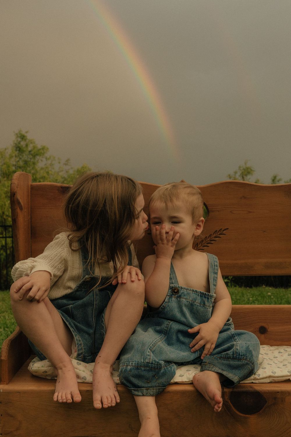 Two people in denim overalls sit on a wooden bench with a rainbow visible in the cloudy sky behind them.