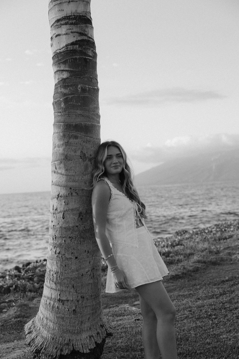 Black and white photo series of a person posing by a palm tree trunk on a beach with ocean views at sunset.