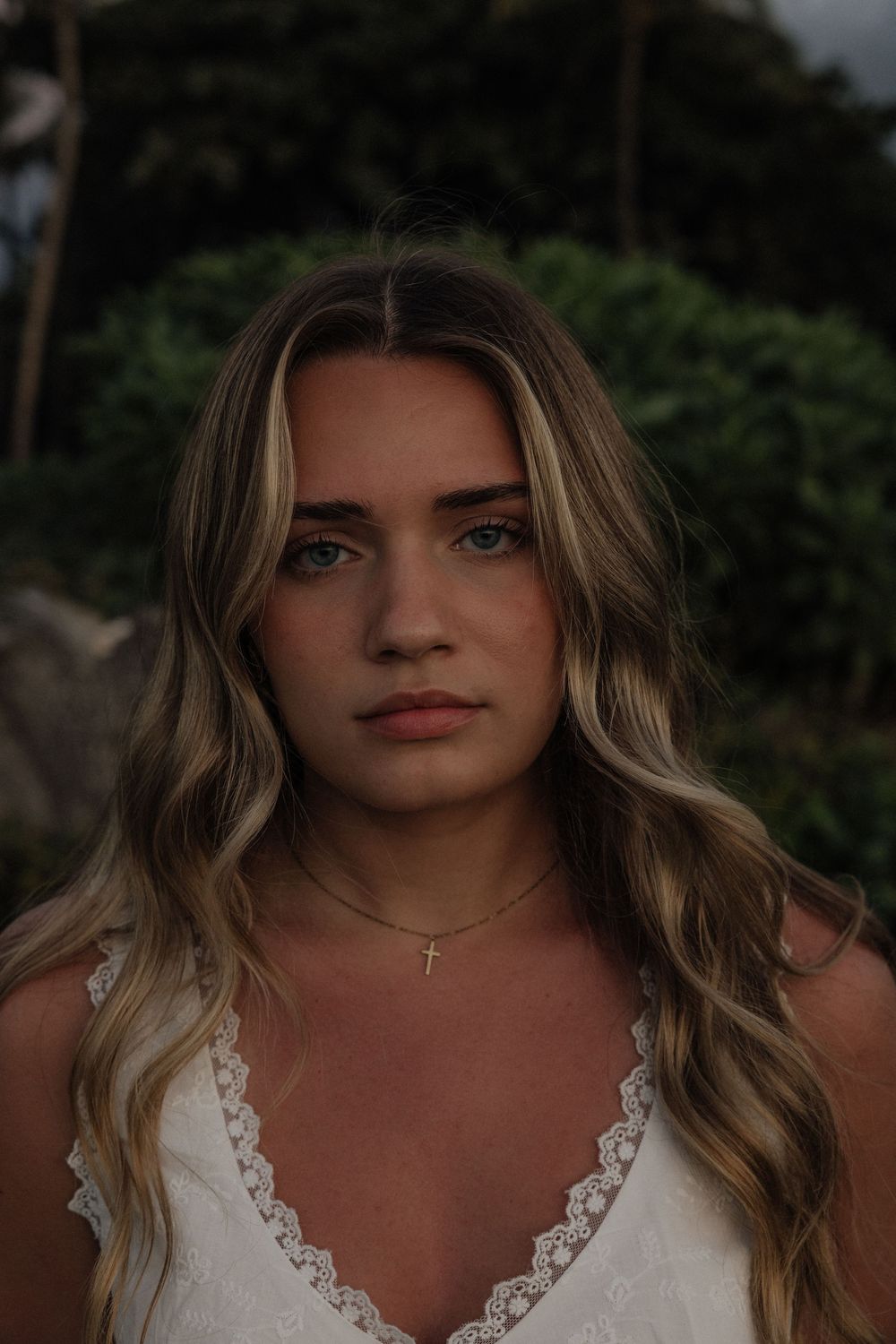 Portrait of a woman with blonde wavy hair wearing a white lace dress against a dark outdoor background.